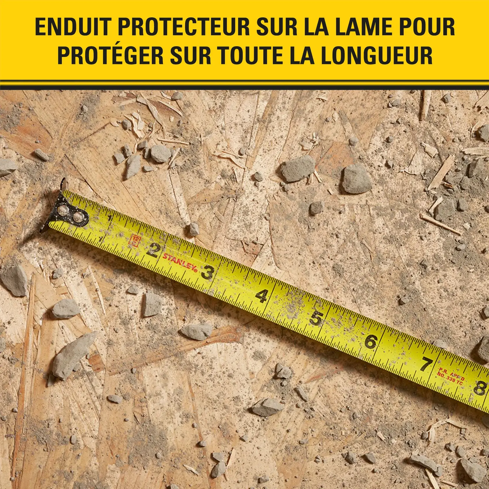 Un ruban à mesurer jaune de la marque Stanley est disposé en diagonale sur une surface en bois rugueuse et poussiéreuse avec des débris. Le ruban montre des marques de mesure et le texte en français au-dessus indique qu'il possède un enduit protecteur sur la lame pour protéger sur toute la longueur.