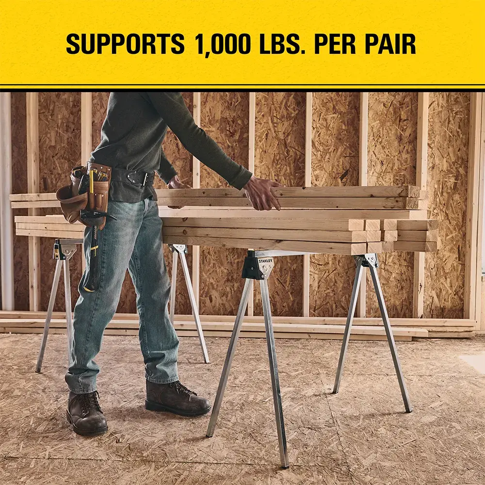 A person standing beside two metal sawhorses holding wooden planks in an unfinished room. A yellow banner above reads, 'Supports 1,000 lbs. per pair.'