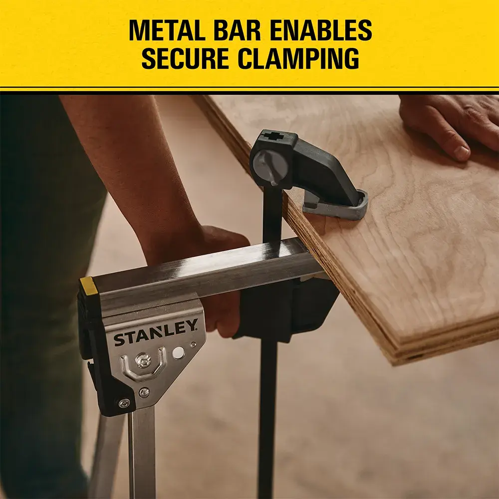 A STANLEY metal bar clamp securely holds a piece of plywood. The image shows a worker using the clamp, with a yellow banner above displaying the text 'Metal bar enables secure clamping.'