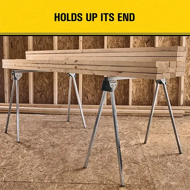 A pair of Stanley STST11154 metal sawhorses holding wooden planks in a workshop setting, with a yellow sign above that reads 'HOLDS UP ITS END'.