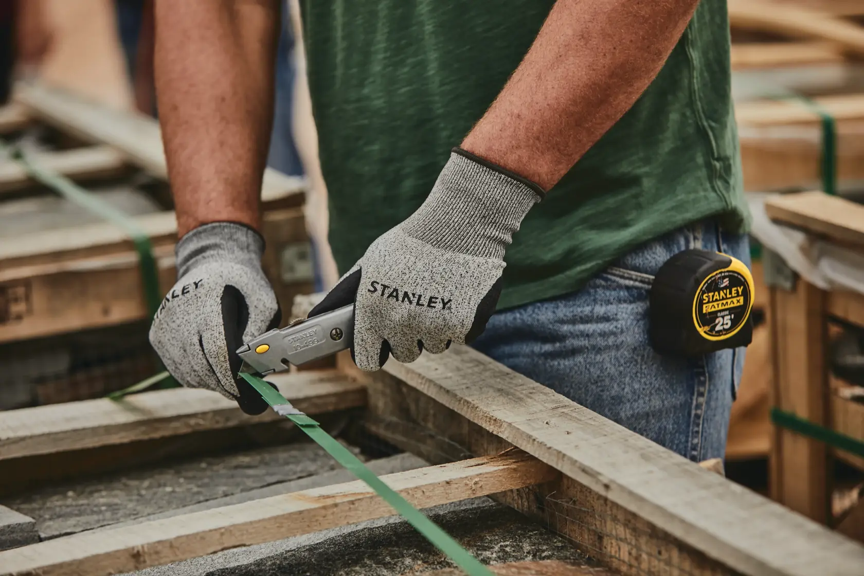 Stanley 10-499 couteau utilitaire en utilisation, ouvrier avec gants coupant une sangle verte sur une palette en bois.