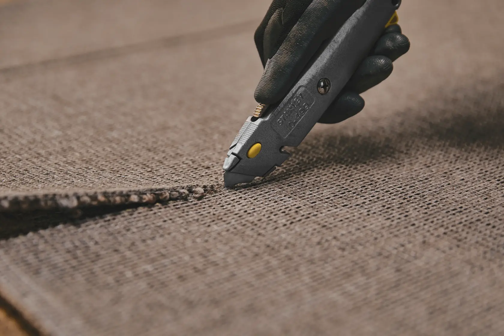 Photographie du couteau utilitaire STANLEY 10-499 découpant un tapis marron, vue rapprochée de la lame en action.