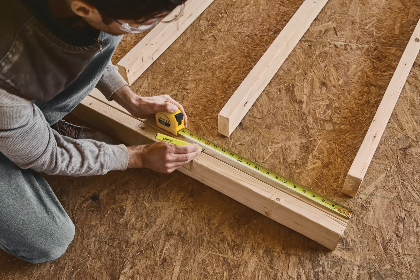 A person measures a wooden beam using a yellow Stanley tape measure on a workshop floor with other wooden pieces nearby.