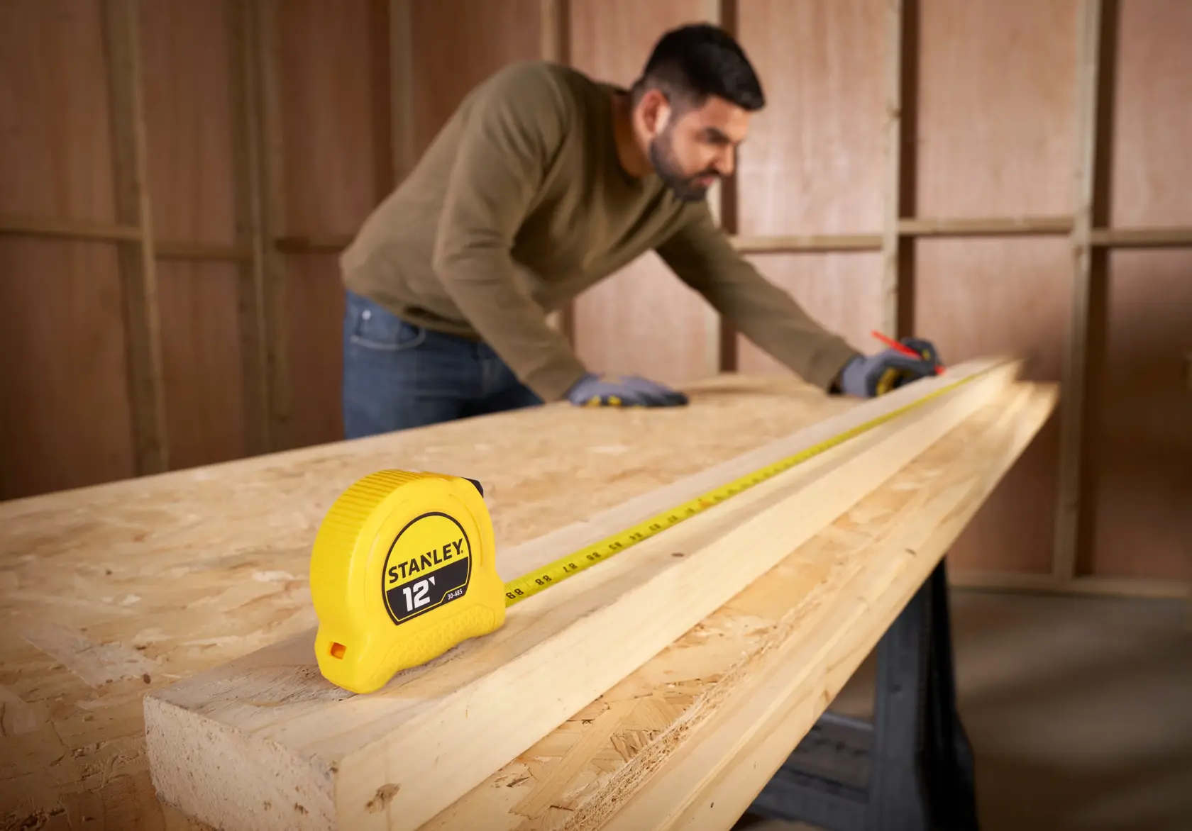 A yellow Stanley 12-foot tape measure is placed on a wooden workbench, with a person in gloves measuring a piece of wood in the background. The scene is set inside a wooden workshop.