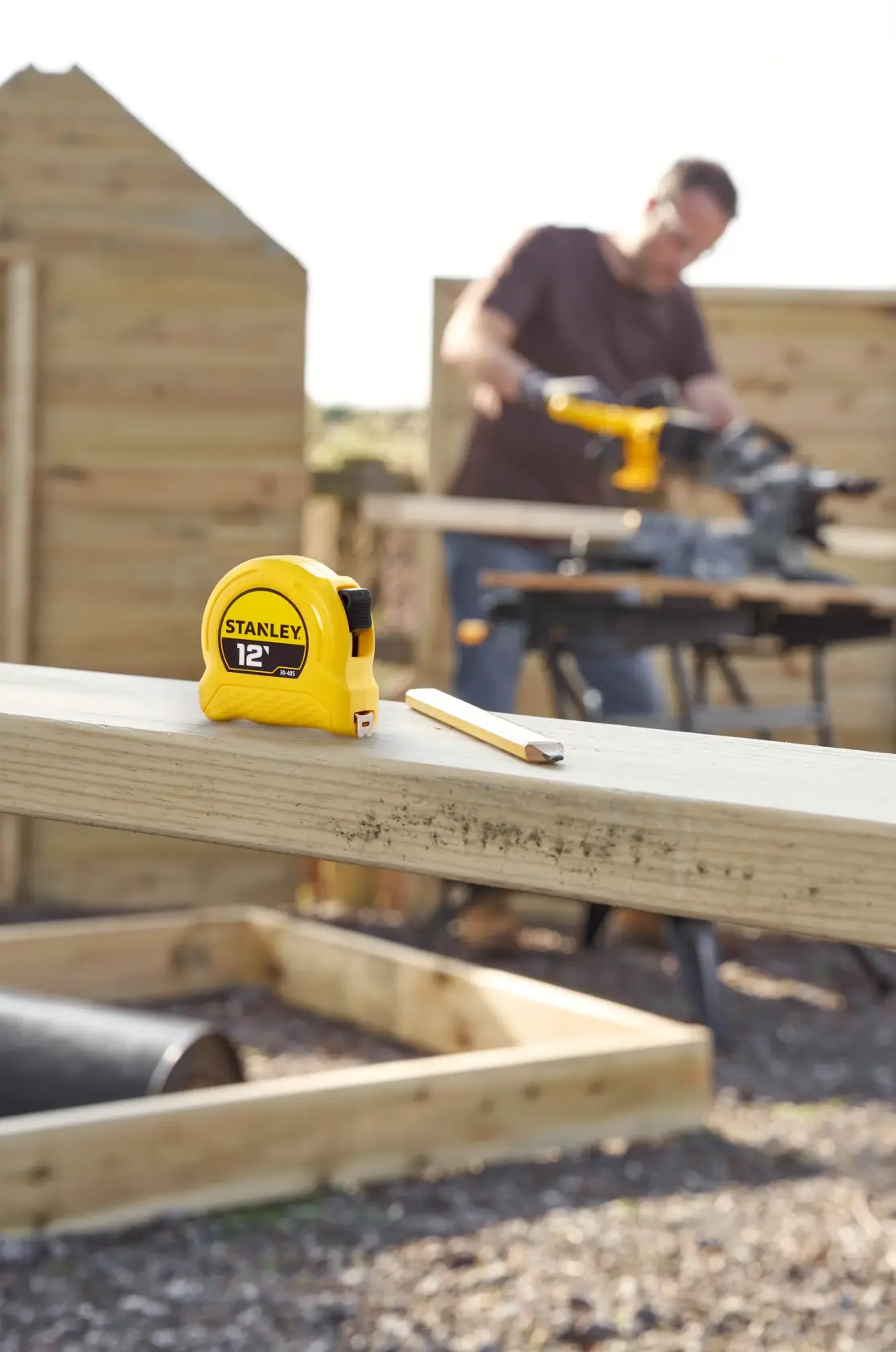 A yellow Stanley 12-foot tape measure and a pencil are placed on a wooden beam at an outdoor construction site. In the background, there is a blurred person working with a power tool, surrounded by wooden structures.