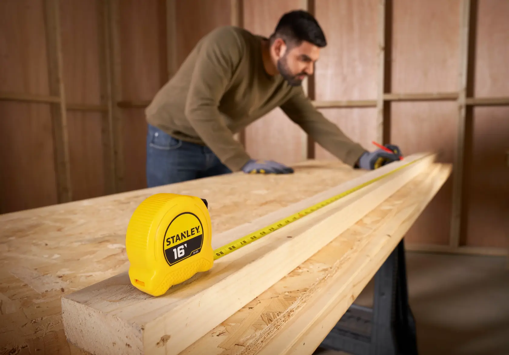 A Stanley 16-foot yellow tape measure is placed on a wooden board. In the background, a person wearing gloves is measuring and marking the board inside a wooden workshop.