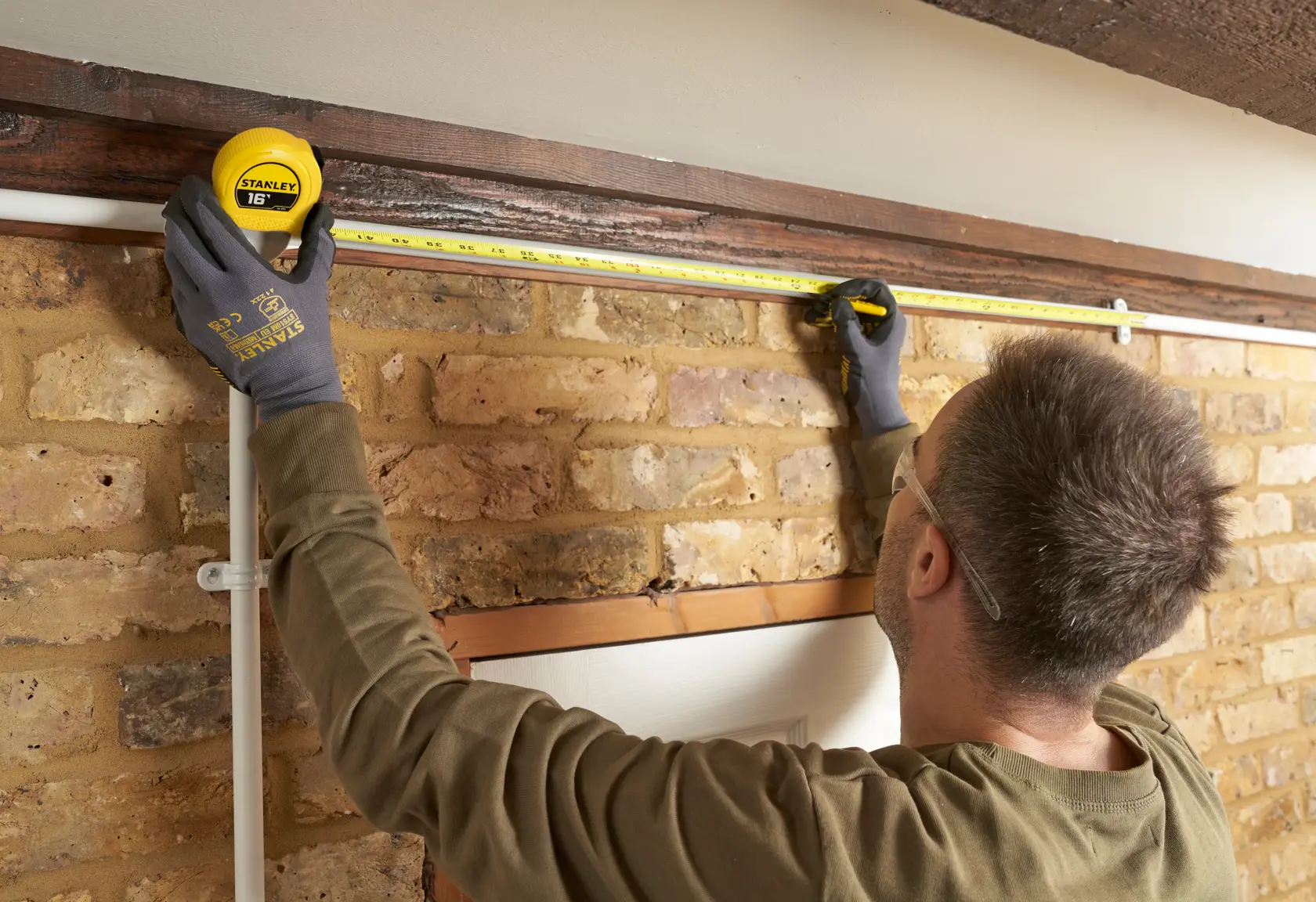 A person wearing protective gloves and safety glasses uses a yellow Stanley tape measure to measure the width above a door frame on a brick wall.