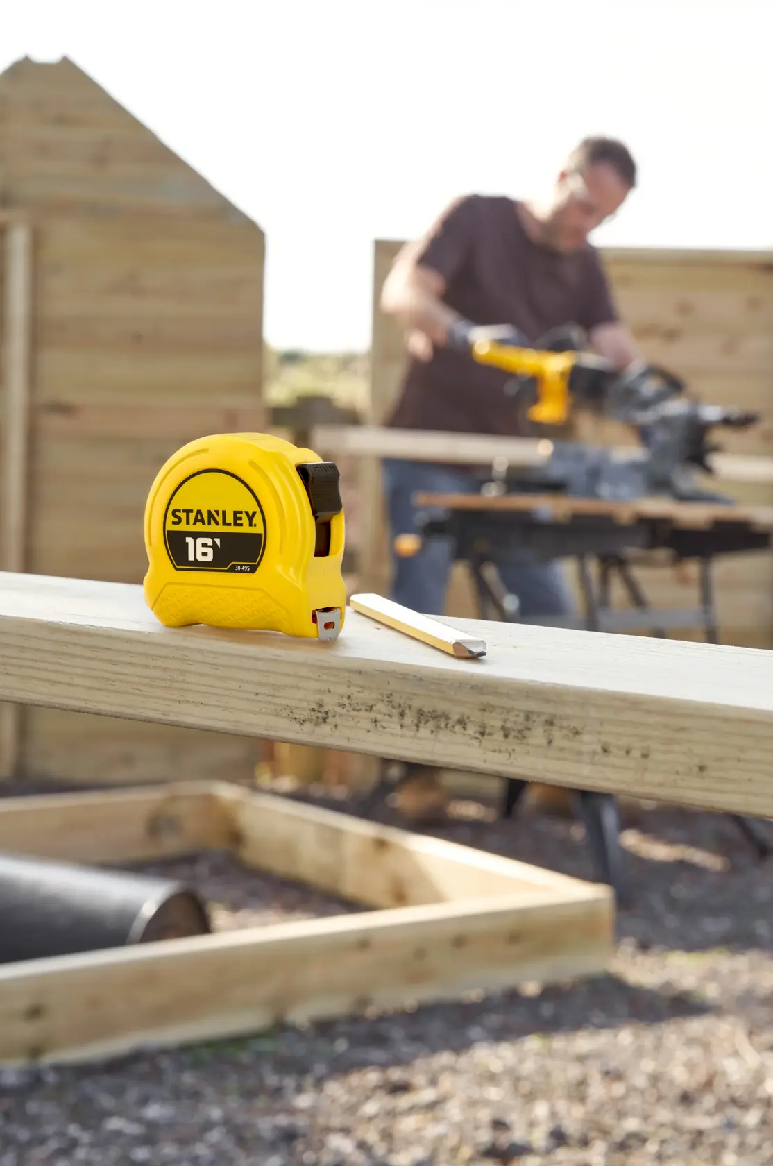 A yellow STANLEY 16-foot tape measure and a pencil are placed on a wooden beam in an outdoor construction setting. In the background, a person is using a power tool with wooden structures and boards around.