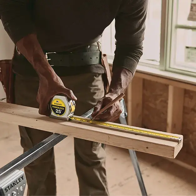 A person measuring a piece of wood with a Stanley tape measure on a workbench in a construction setting.
