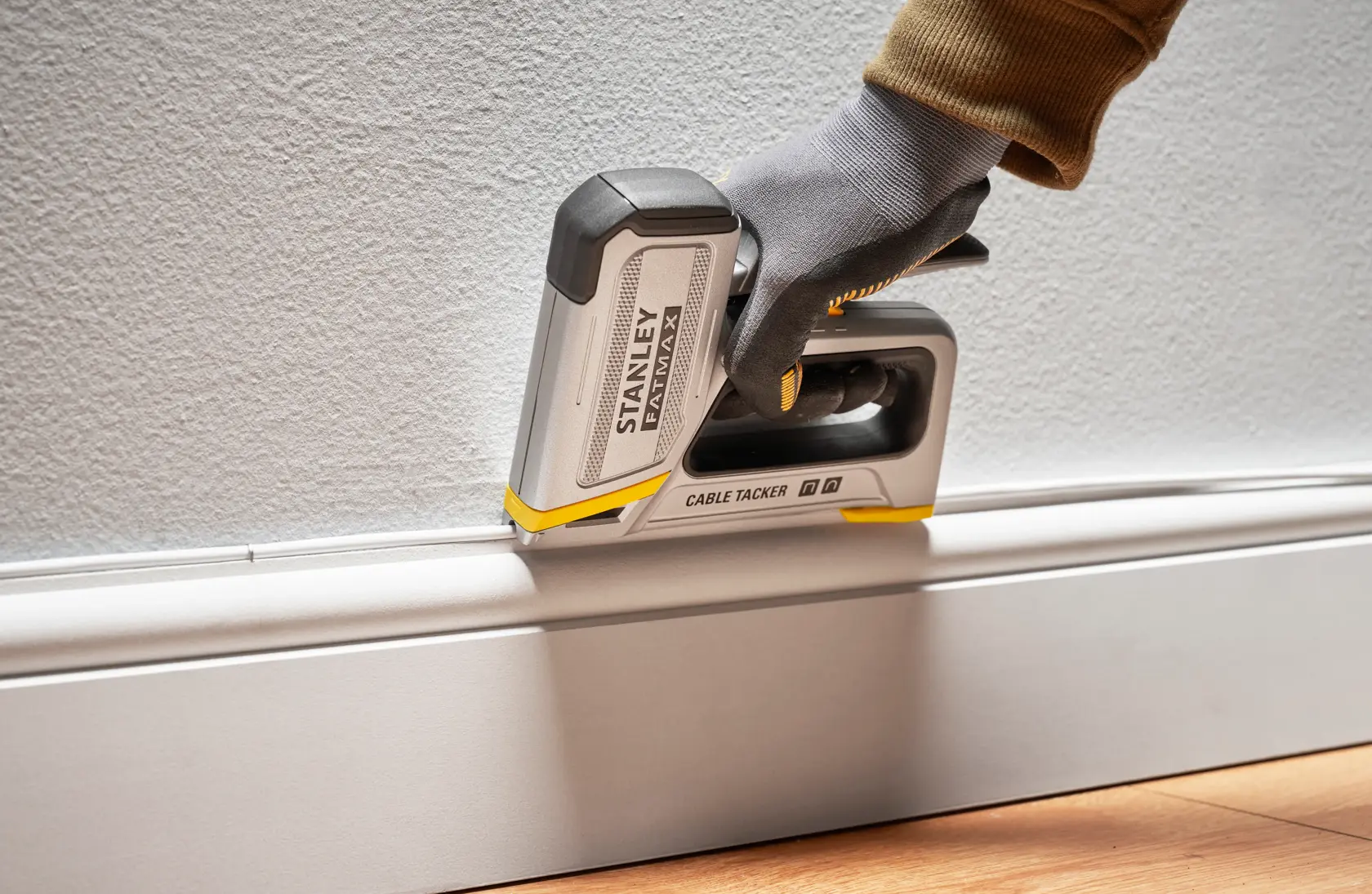 Photo of STANLEY FMHT70100 cable tacker securing wire along baseboard, side angle, gloved hand operating tool.