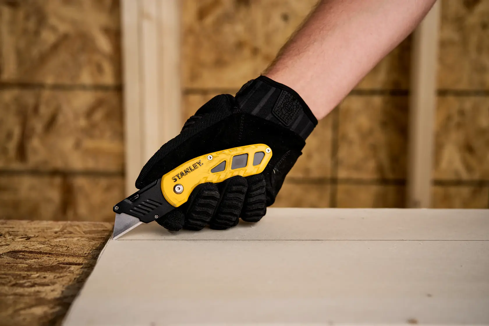 STANLEY STHT10424 folding utility knife, yellow and black, in gloved hand cutting drywall, close-up.