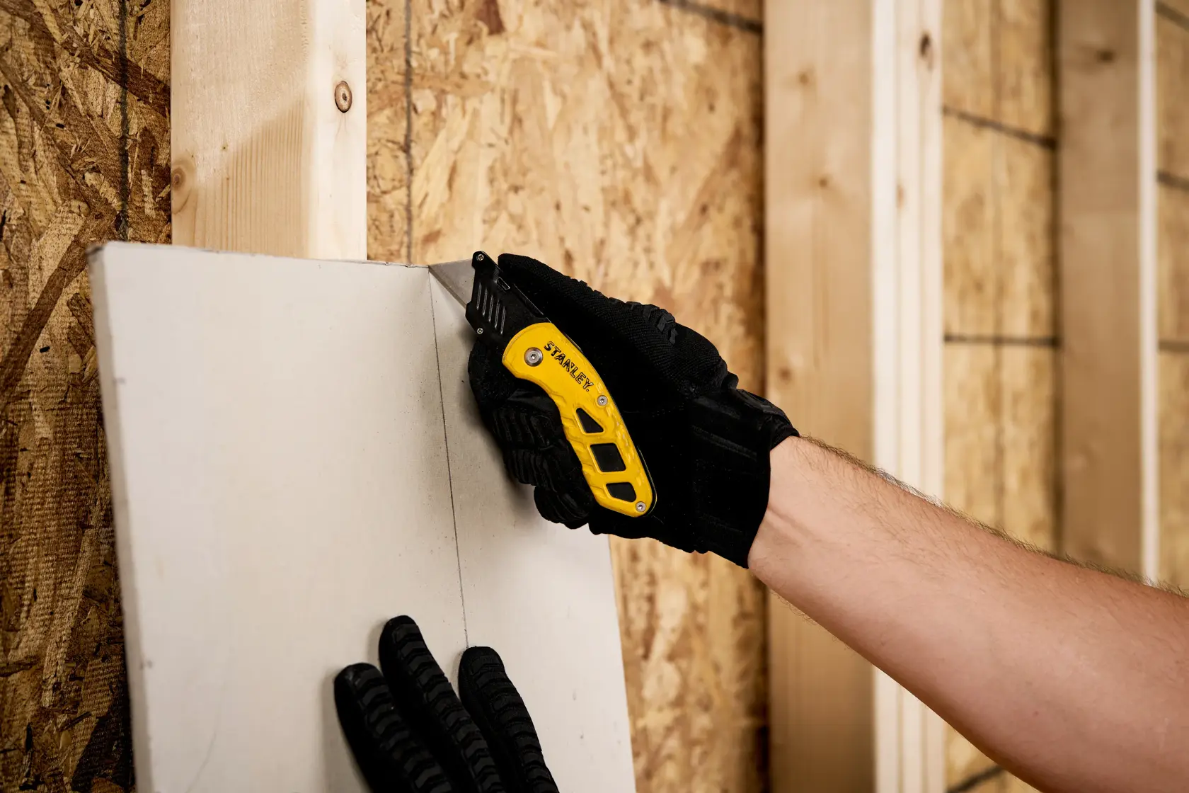 Photo of STANLEY STHT10424 utility knife cutting drywall, held by gloved hand in construction setting.