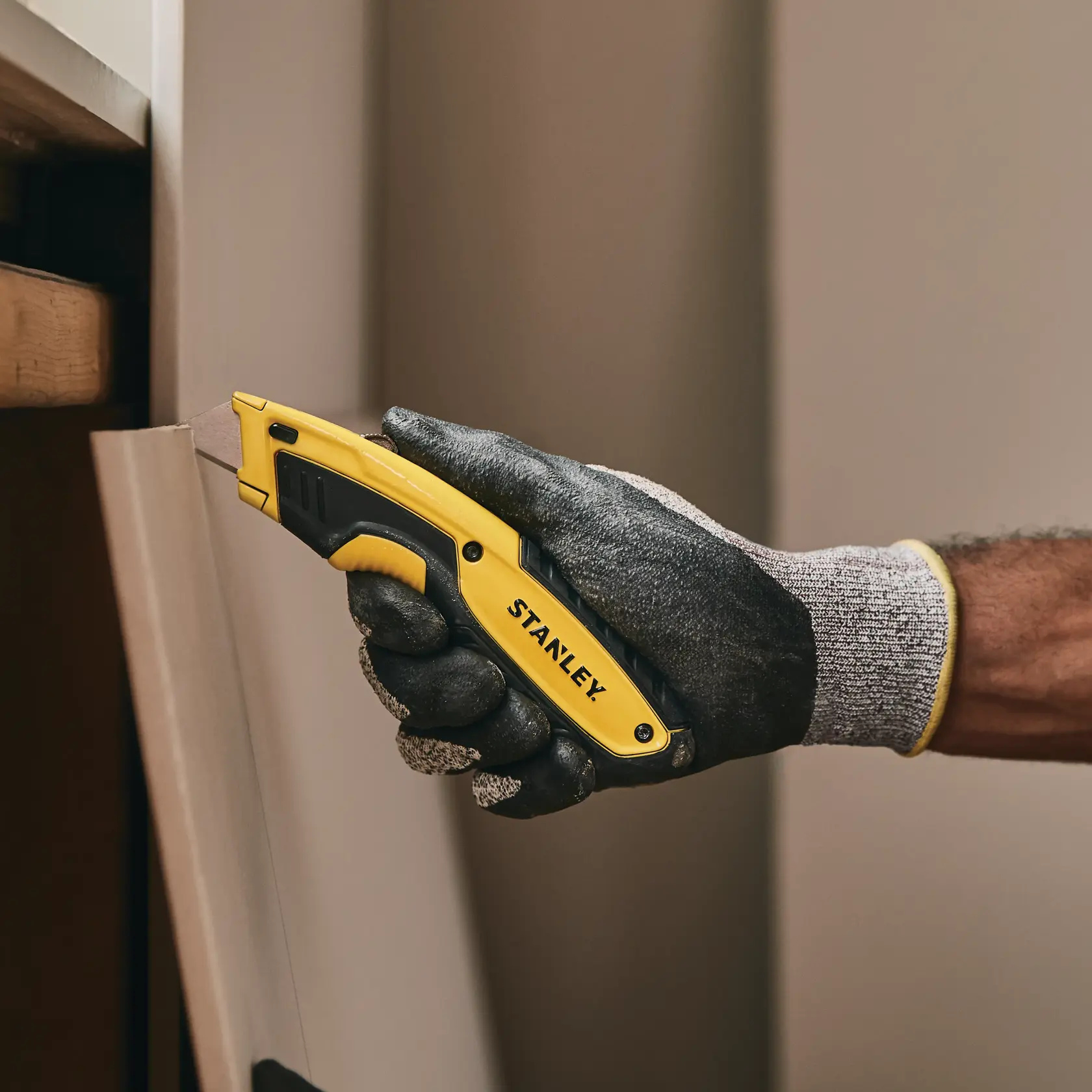 STANLEY STHT10479 utility knife with yellow and black handle, gloved hand cutting wood panel.
