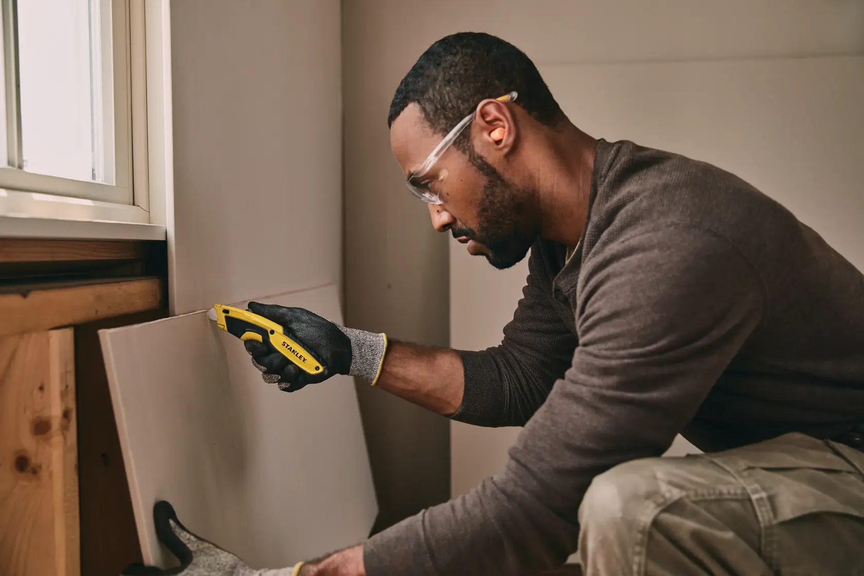 STANLEY STHT10479 utility knife held by a gloved hand cutting drywall in an indoor workspace.