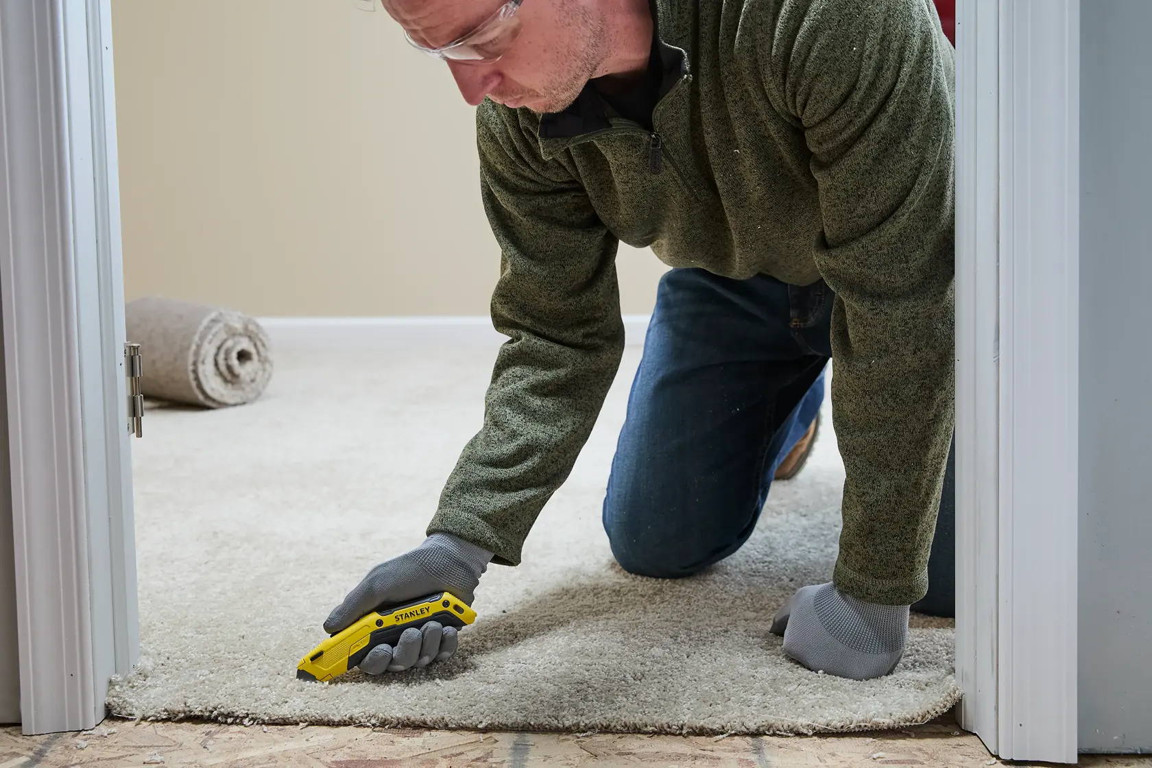 A person wearing gloves, a green sweater, and jeans is kneeling on a carpet, using a yellow Stanley utility knife to cut the carpet near a doorway. A rolled carpet is visible in the background.
