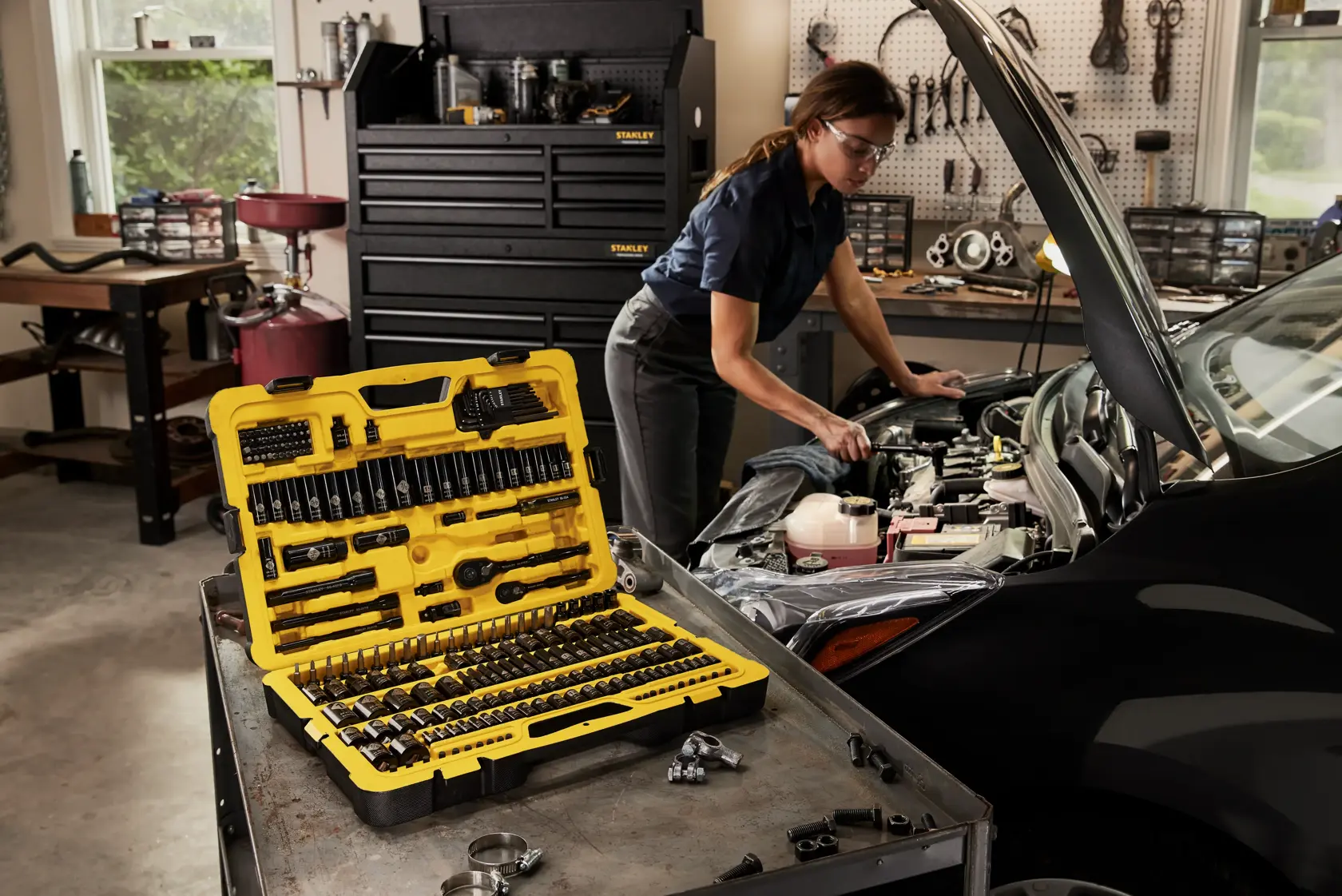 STANLEY STMT75064 mechanic tool set open on a workbench beside a car in a garage, showing assorted tools in a yellow case.