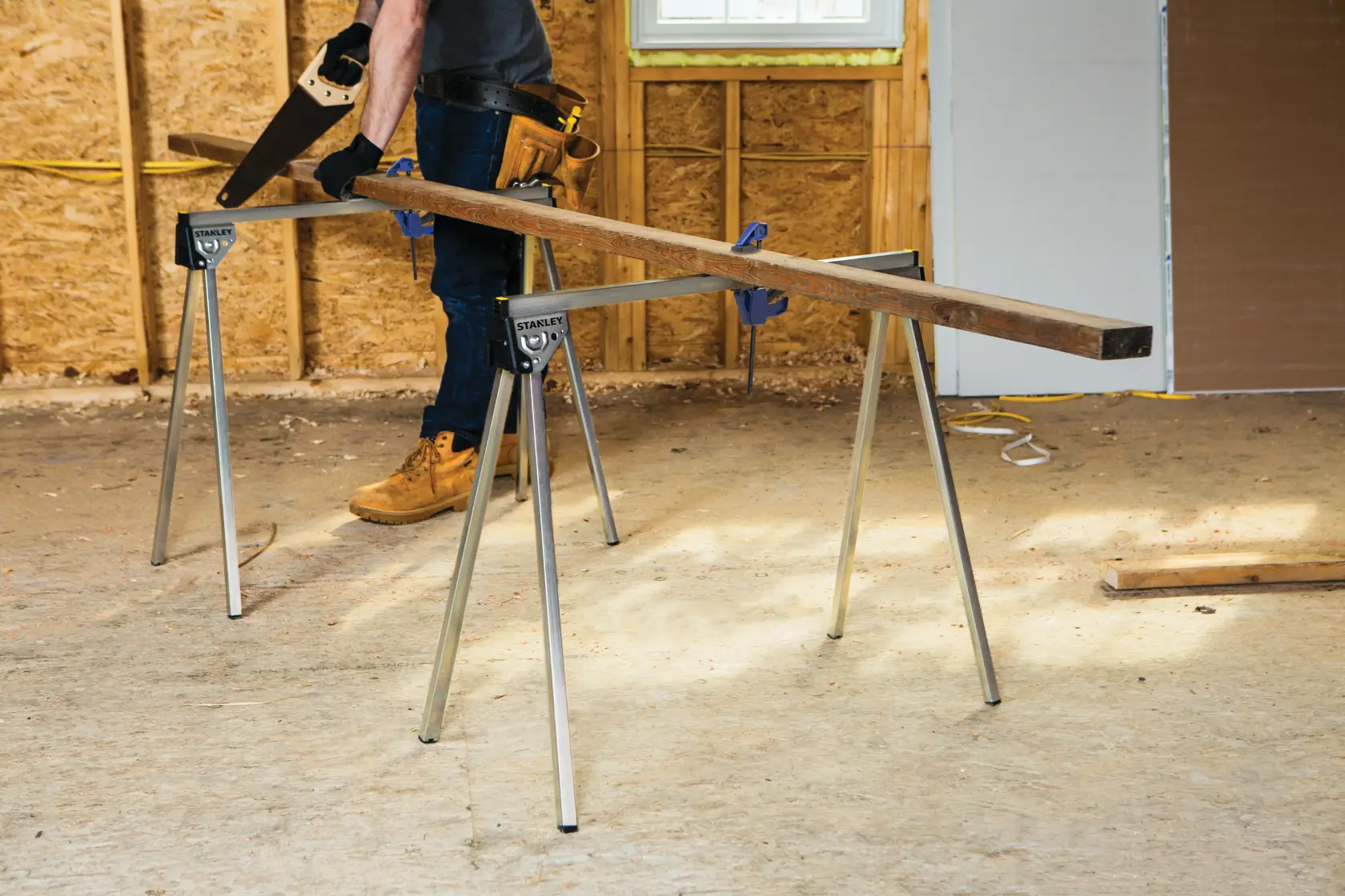 A person standing beside two metal sawhorses holding wooden planks in an unfinished room. A yellow banner above reads, 'Supports 1,000 lbs. per pair.'