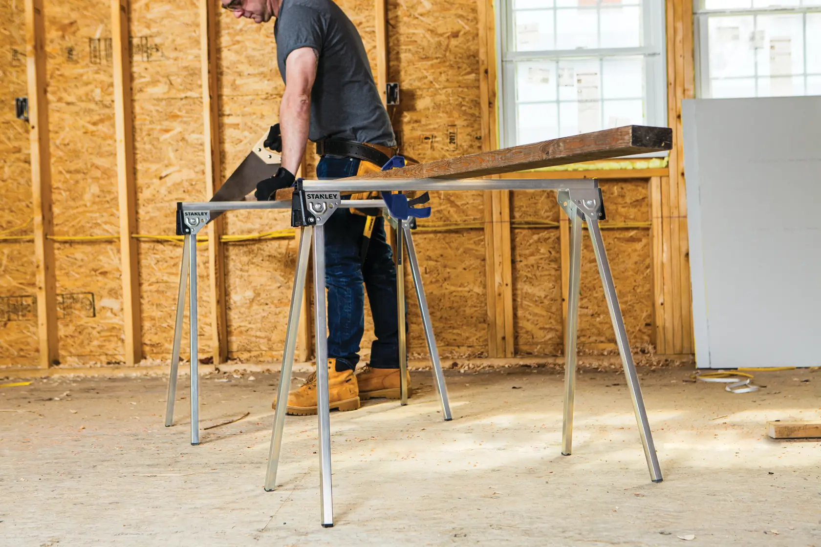 A STANLEY metal bar clamp securely holds a piece of plywood. The image shows a worker using the clamp, with a yellow banner above displaying the text 'Metal bar enables secure clamping.'